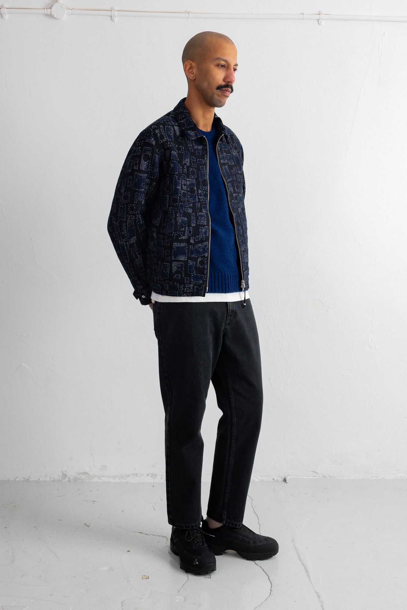 A man wearing a dark patterned bomber jacket over a blue shirt, black pants, and black sneakers, standing against a white studio wall.