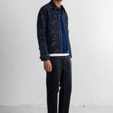A man wearing a dark blue and black patterned button-up shirt jacket over a blue t-shirt, black jeans, and black sneakers, standing against a white studio wall.