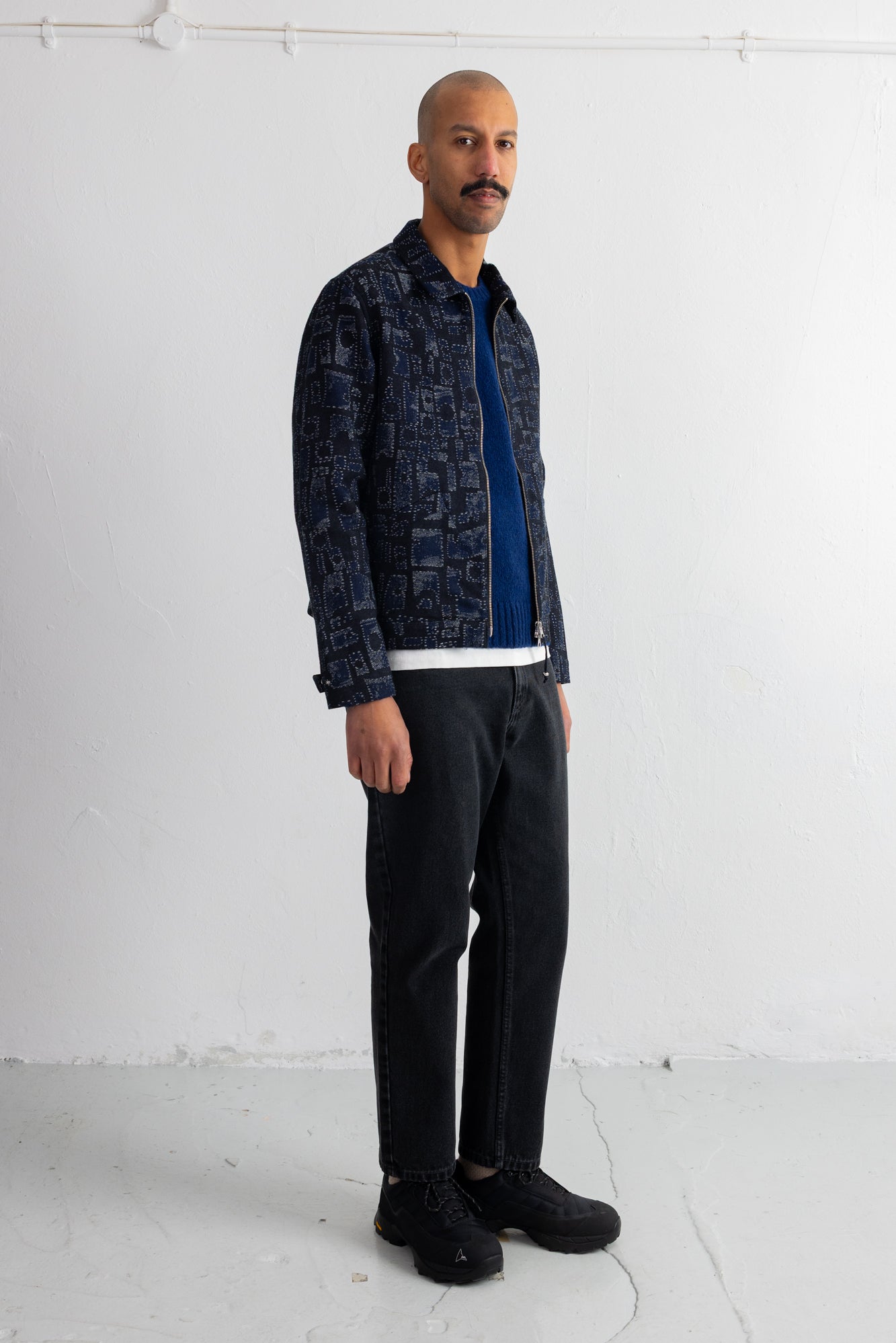 A man wearing a dark blue and black patterned button-up shirt jacket over a blue t-shirt, black jeans, and black sneakers, standing against a white studio wall.