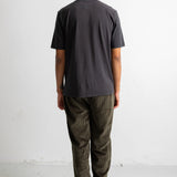 A man viewed from behind wearing a dark charcoal gray t-shirt and olive green pants, standing against a plain white studio wall.