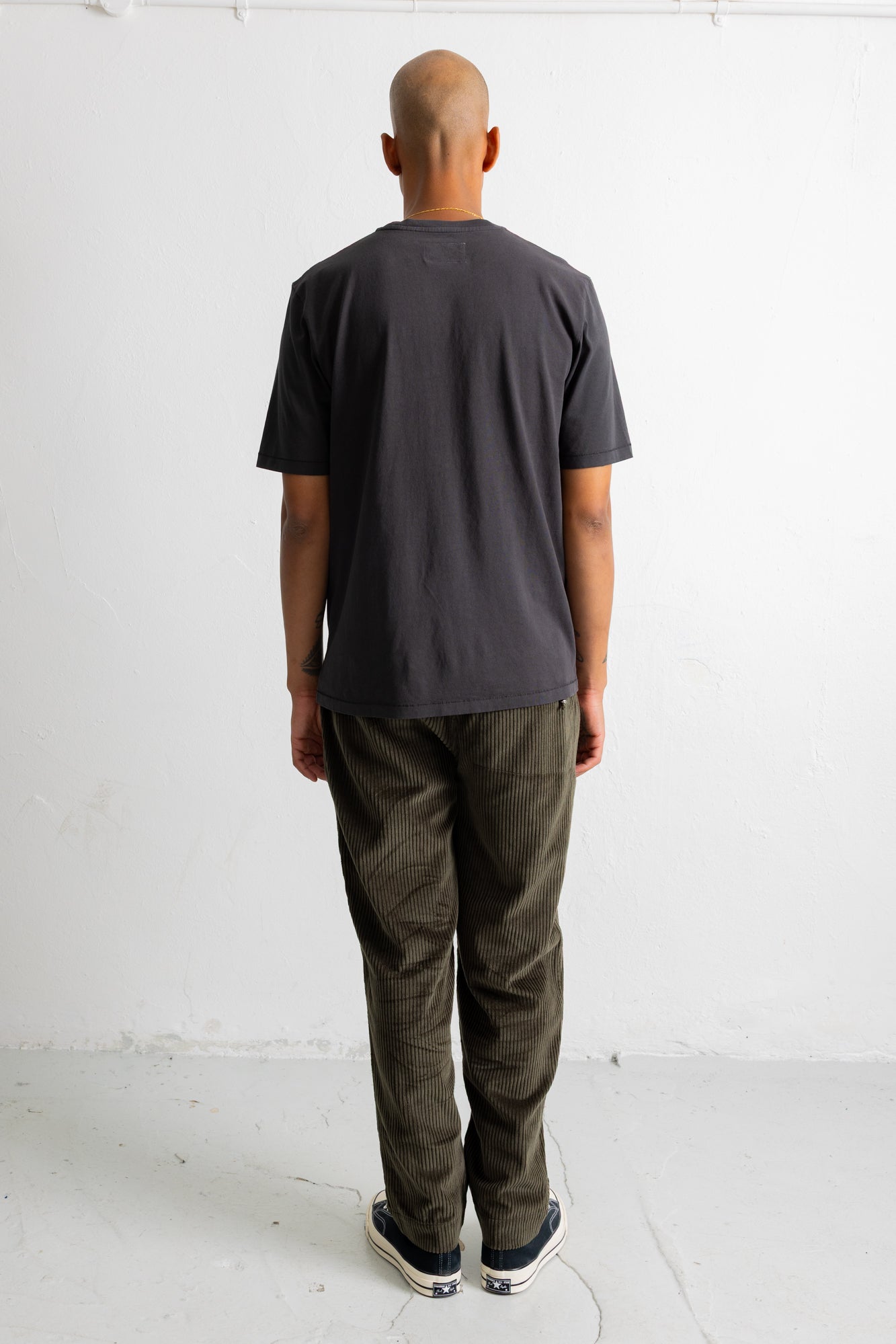 A man viewed from behind wearing a dark charcoal gray t-shirt and olive green pants, standing against a plain white studio wall.