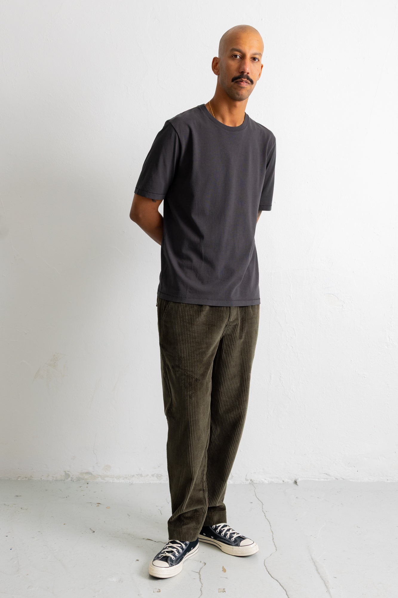 A man wearing a dark charcoal gray crewneck t-shirt, olive green corduroy pants, and black canvas sneakers with white soles, standing against a white studio wall.