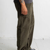 A person wearing wide-leg olive green corduroy pants, a dark gray t-shirt, and black canvas sneakers, standing against a white textured wall.