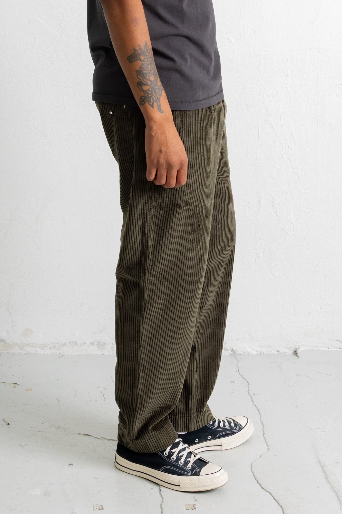 A person wearing wide-leg olive green corduroy pants, a dark gray t-shirt, and black canvas sneakers, standing against a white textured wall.