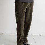 A person wearing dark olive corduroy pants, a dark charcoal t-shirt, and navy blue canvas sneakers with white soles, standing against a white wall.