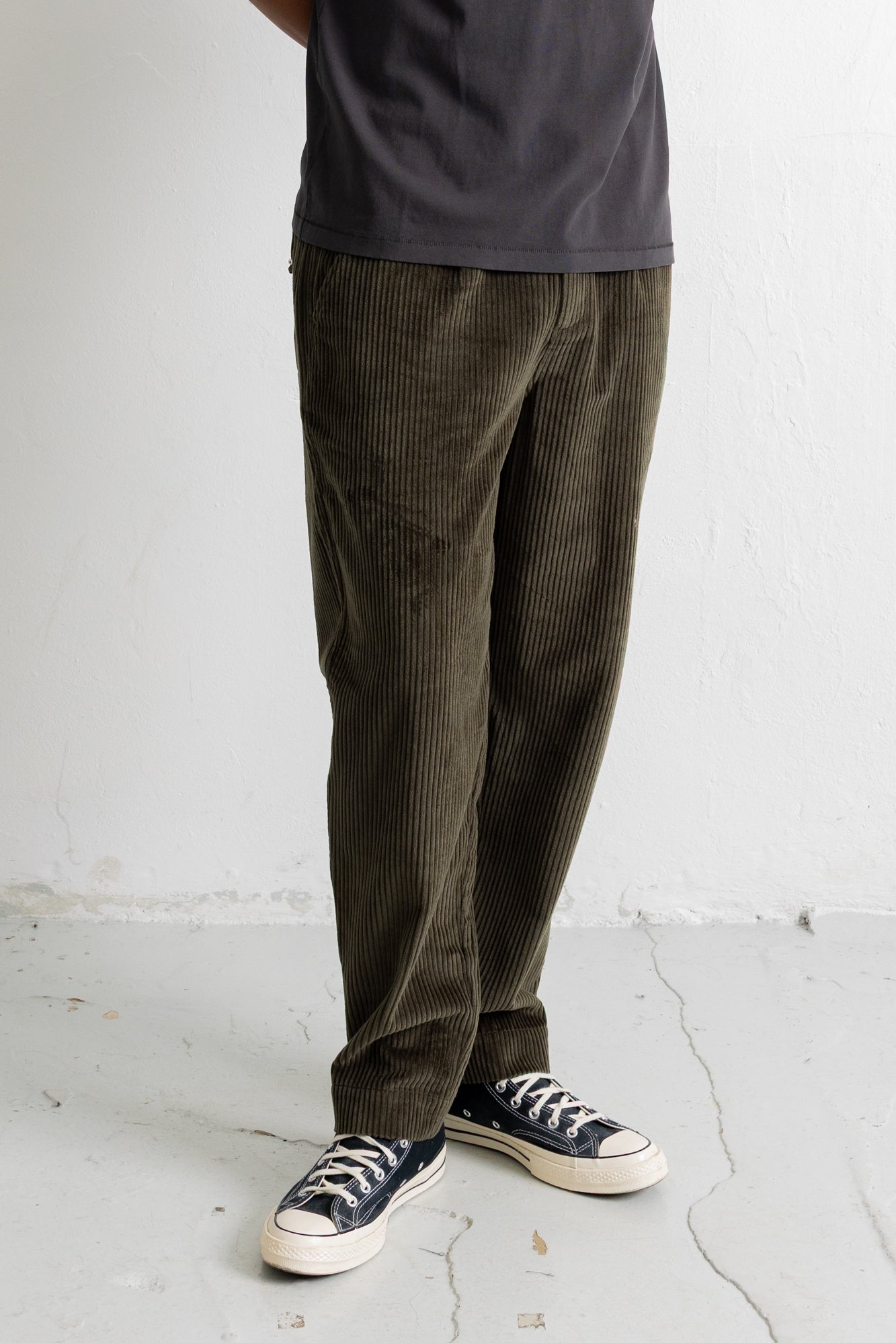 A person wearing dark olive corduroy pants, a dark charcoal t-shirt, and navy blue canvas sneakers with white soles, standing against a white wall.