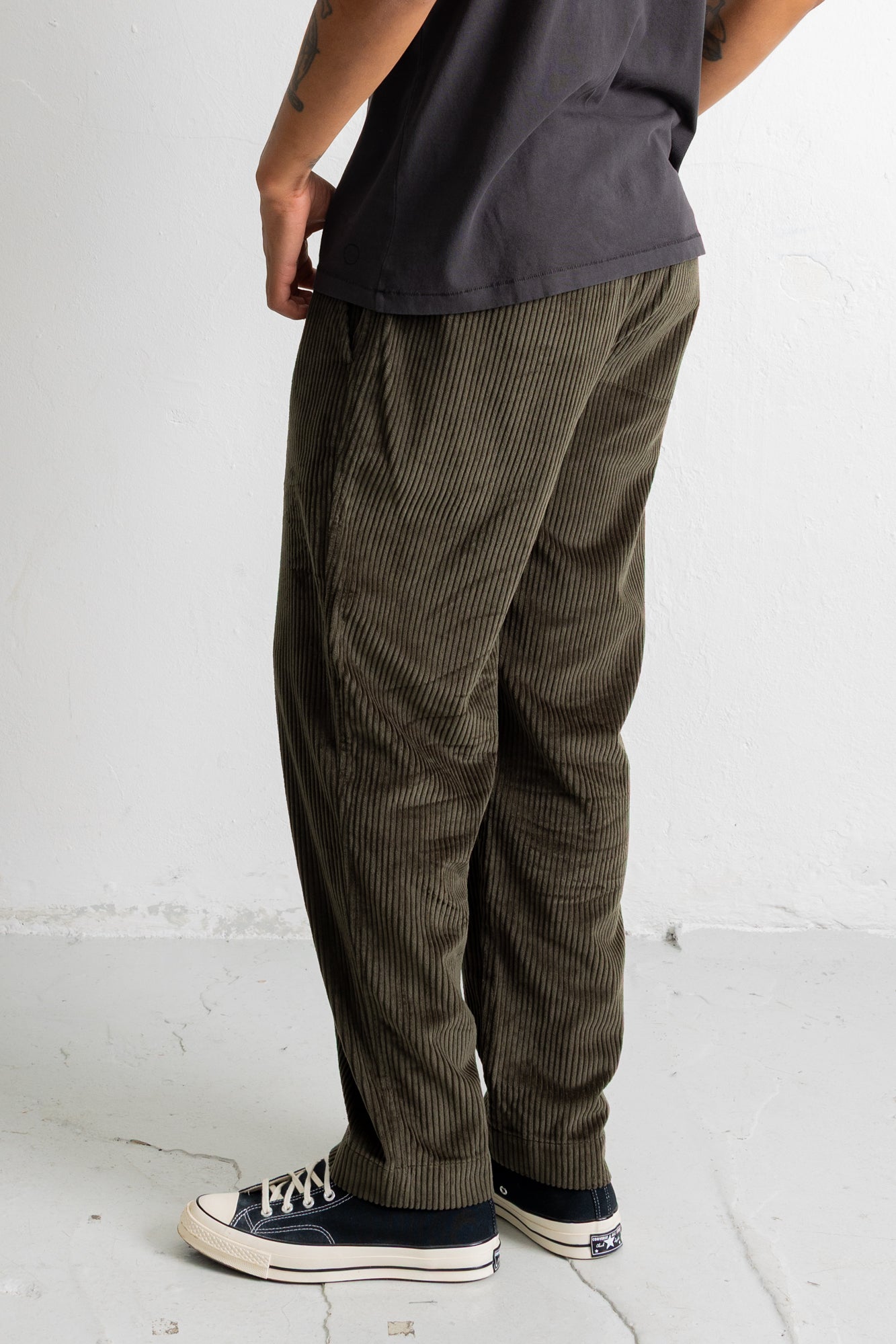 A person shown from the waist down wearing dark olive corduroy pants, a faded black t-shirt, and navy blue canvas sneakers against a white studio wall.