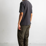 A man shown from the back wearing a dark charcoal gray t-shirt, olive green corduroy pants, and black canvas sneakers, standing against a plain white wall.