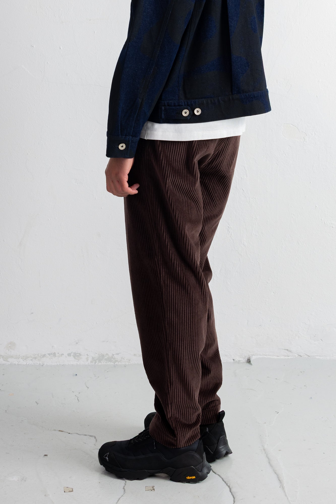 A person shown from the back wearing dark brown corduroy pants, a dark navy jacket, a white undershirt, and black sneakers, standing against a white wall.