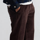 A close-up of a person wearing dark brown corduroy trousers with a hand in the pocket, styled with a white undershirt and a dark patterned jacket against a white wall.