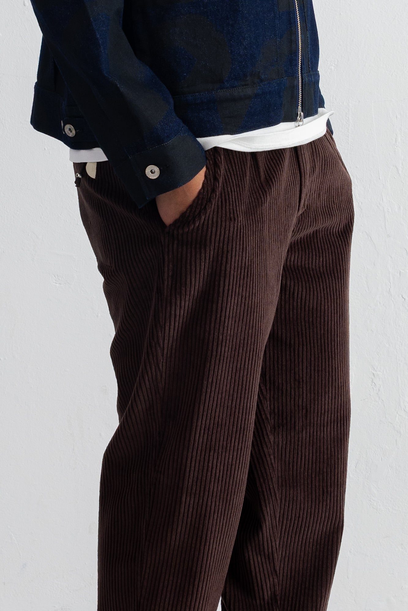 A close-up of a person wearing dark brown corduroy trousers with a hand in the pocket, styled with a white undershirt and a dark patterned jacket against a white wall.
