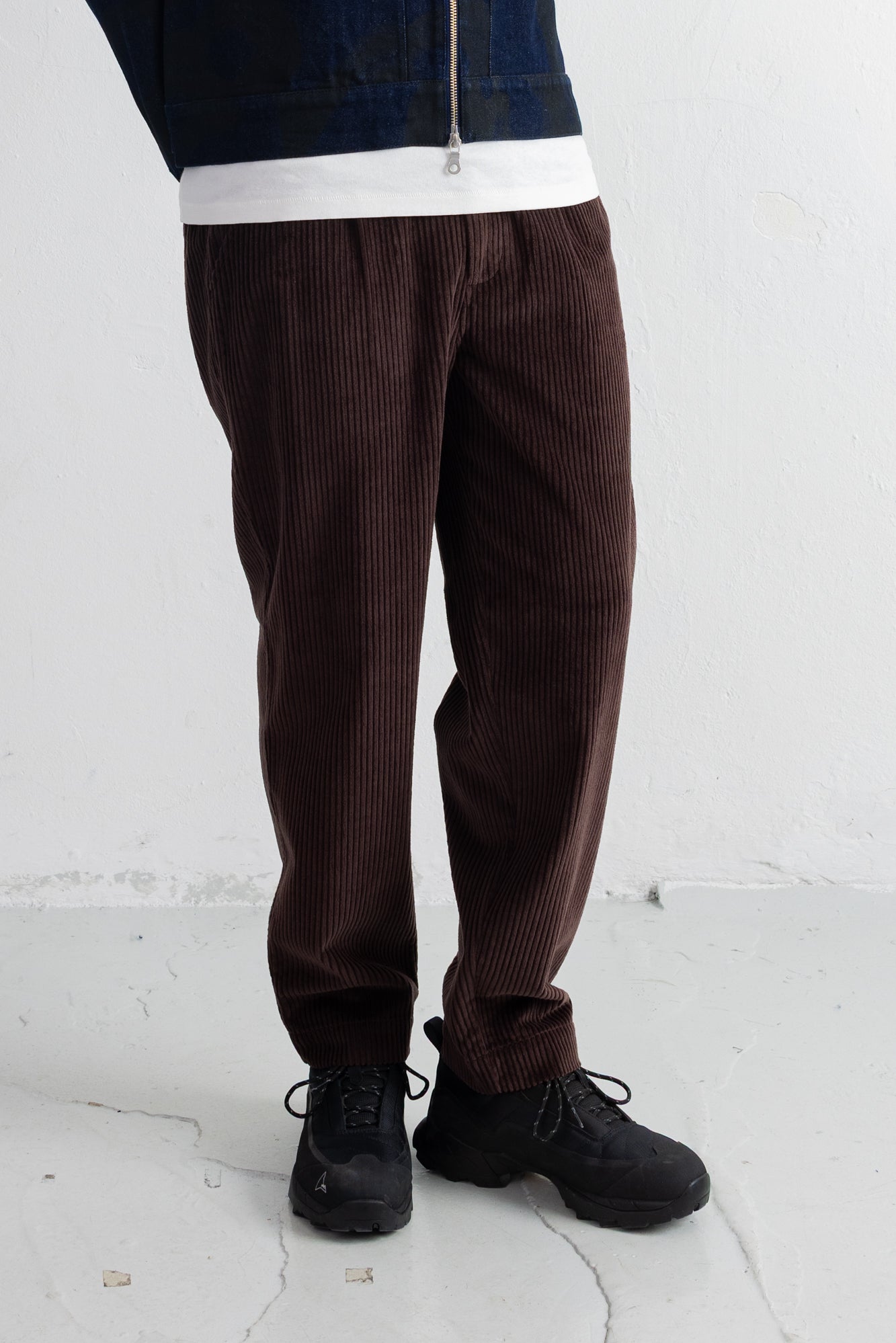 A person wearing dark brown corduroy pants, a dark blue zip-up jacket, a white shirt, and black shoes, standing against a white wall.