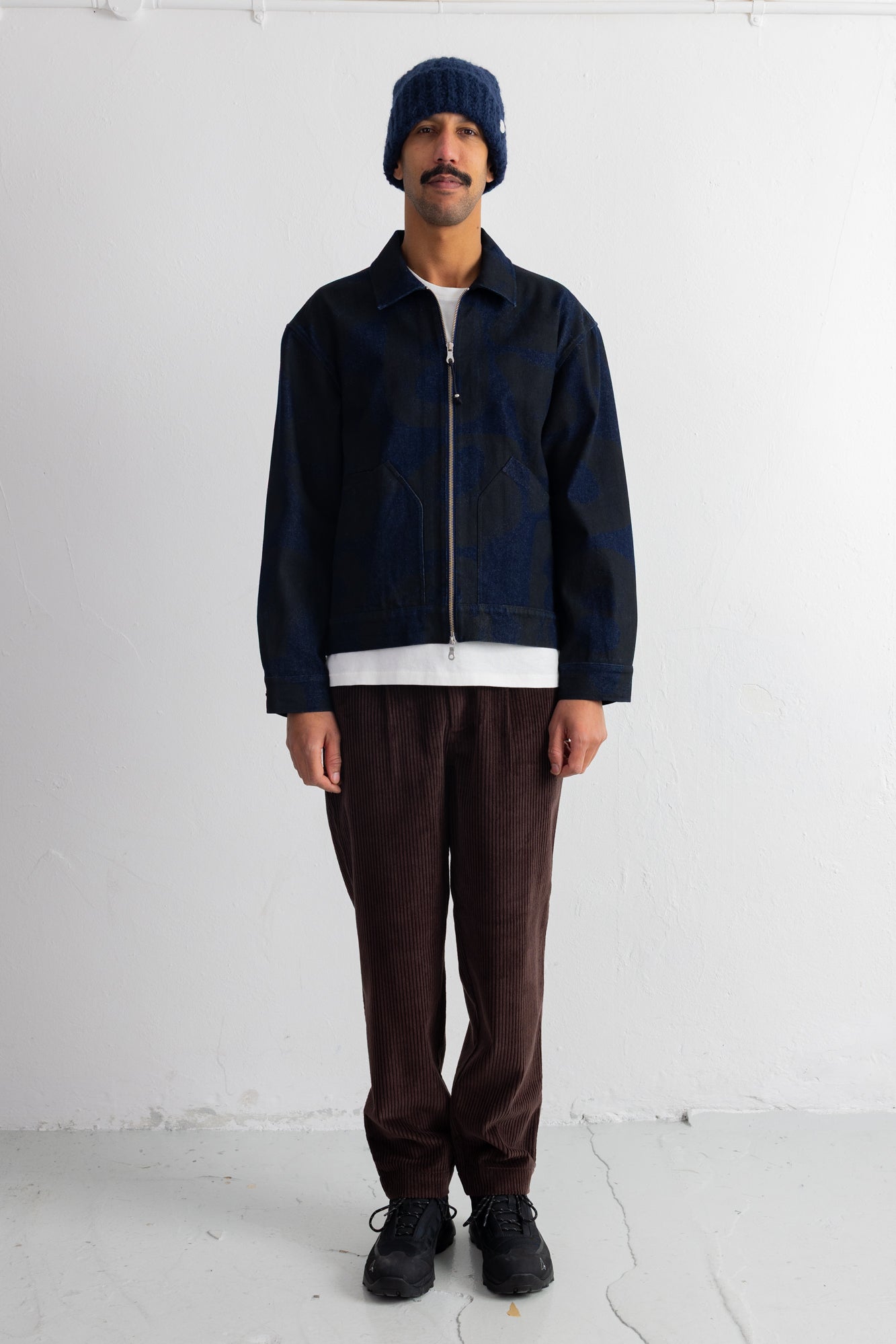 A man wearing a dark navy blue zip-up jacket, dark brown pants, a white t-shirt, black sneakers, and a navy beanie hat, standing against a white studio wall.