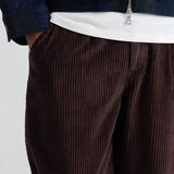Close-up of a person wearing dark brown corduroy trousers, a white t-shirt, and a dark blue zip-up jacket against a plain light background.