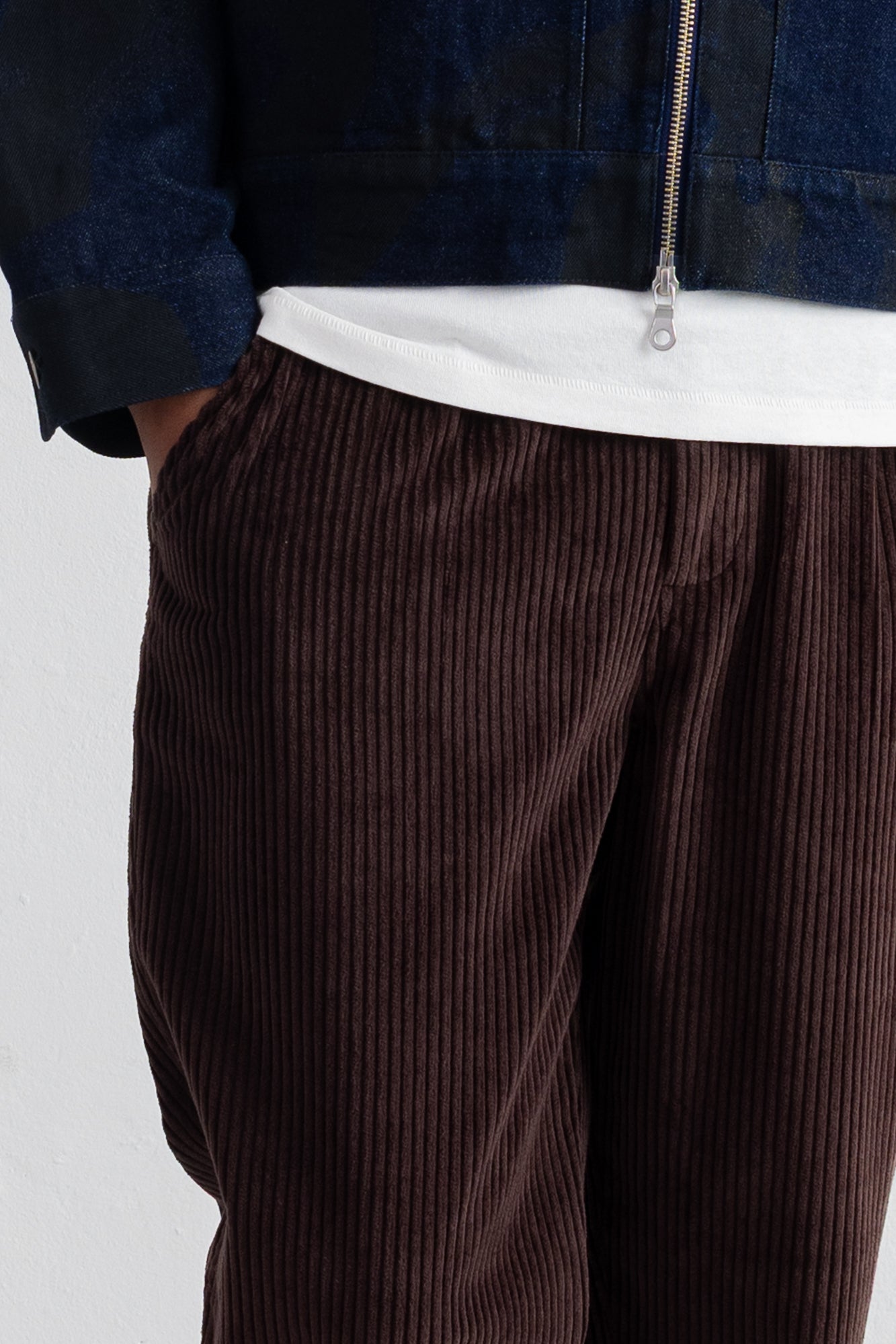 Close-up of a person wearing dark brown corduroy trousers, a white t-shirt, and a dark blue zip-up jacket against a plain light background.