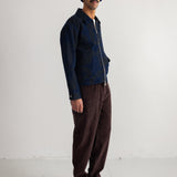 A man wearing a dark blue zip-up jacket with a dark blue knit beanie, brown corduroy pants, and black shoes, standing against a plain white studio wall.