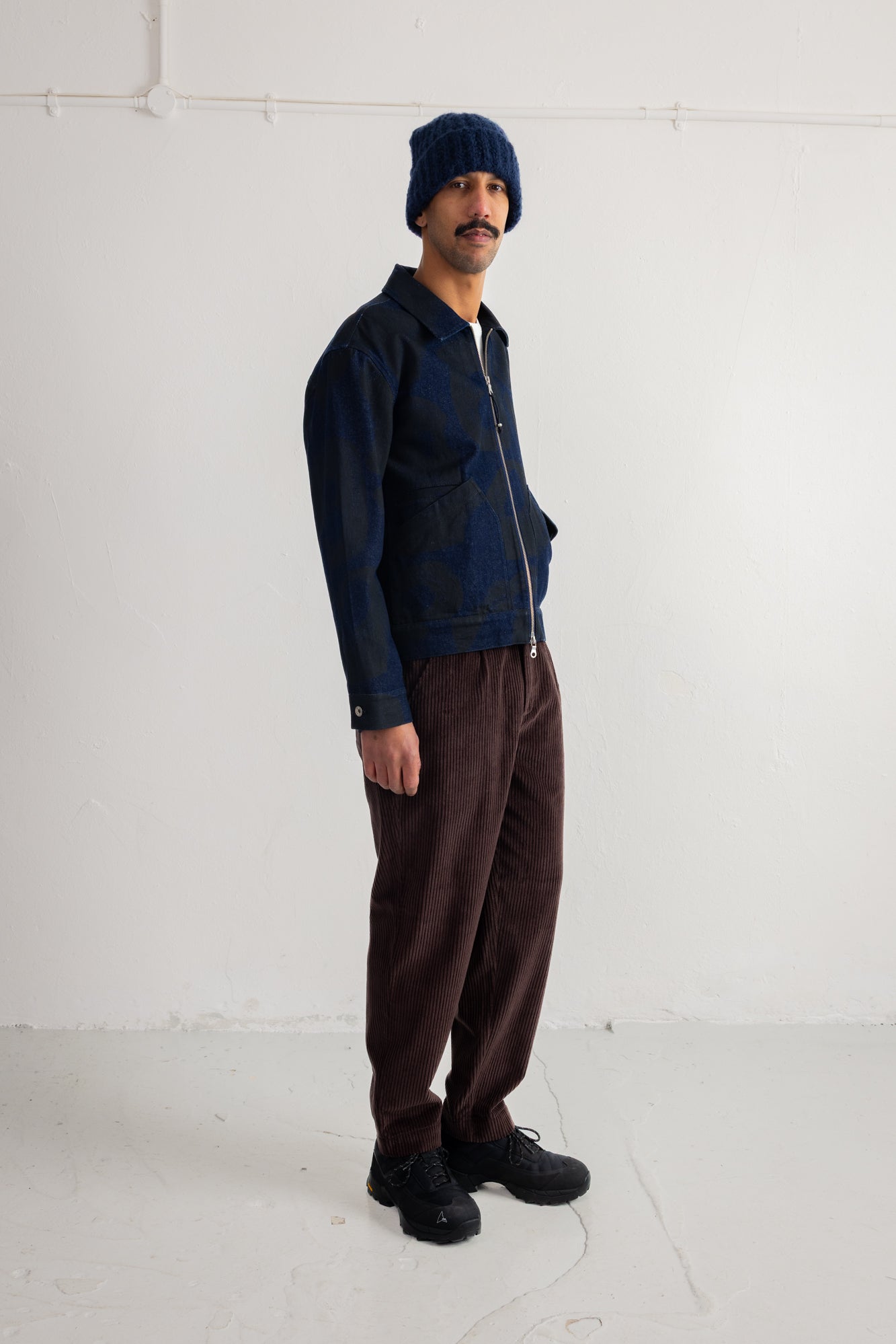 A man wearing a dark blue zip-up jacket with a dark blue knit beanie, brown corduroy pants, and black shoes, standing against a plain white studio wall.