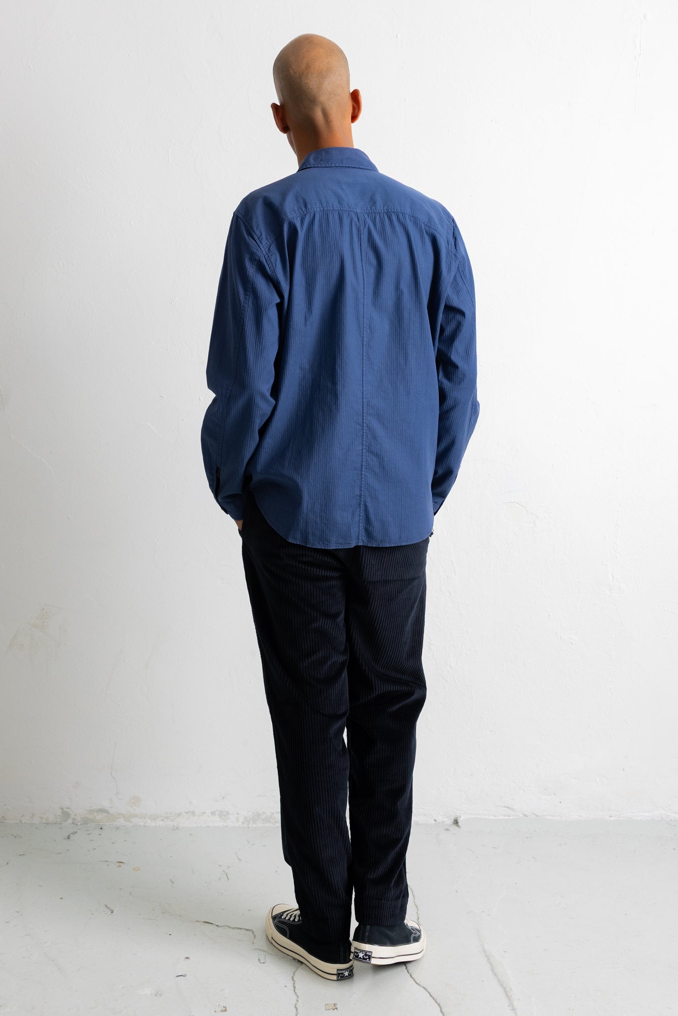A man viewed from behind wearing a blue long-sleeve button-up shirt, dark trousers, and black-and-white sneakers, standing against a plain white wall.