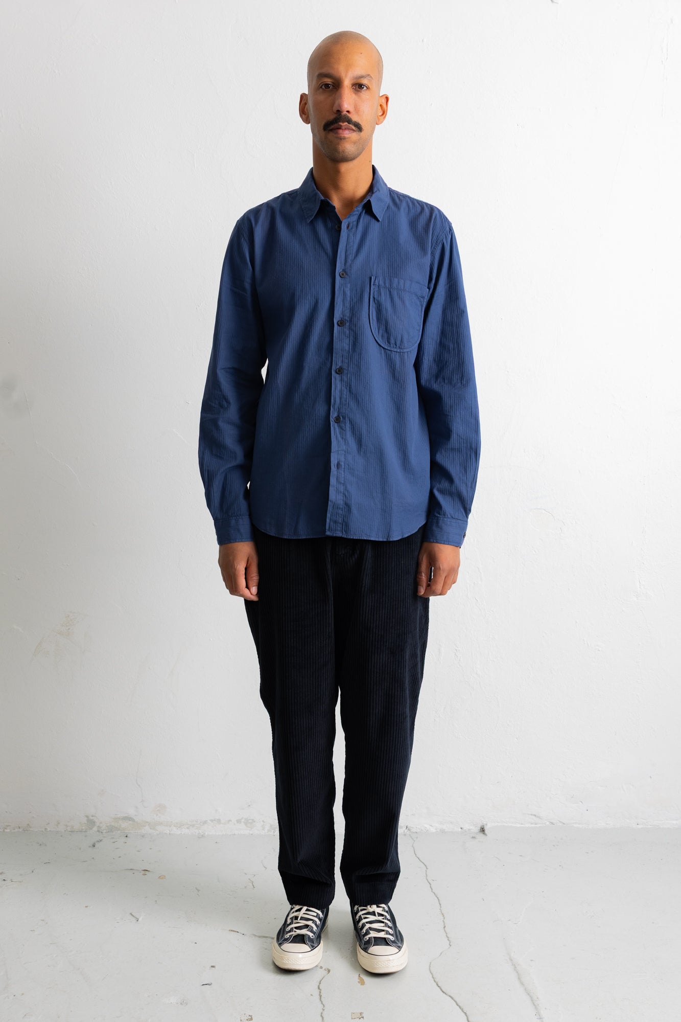 A man wearing a blue long-sleeve button-down shirt, dark pants, and sneakers, standing against a plain white wall.