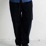A person wearing dark navy corduroy pants, a blue button-up shirt, and black canvas sneakers, standing against a white wall.