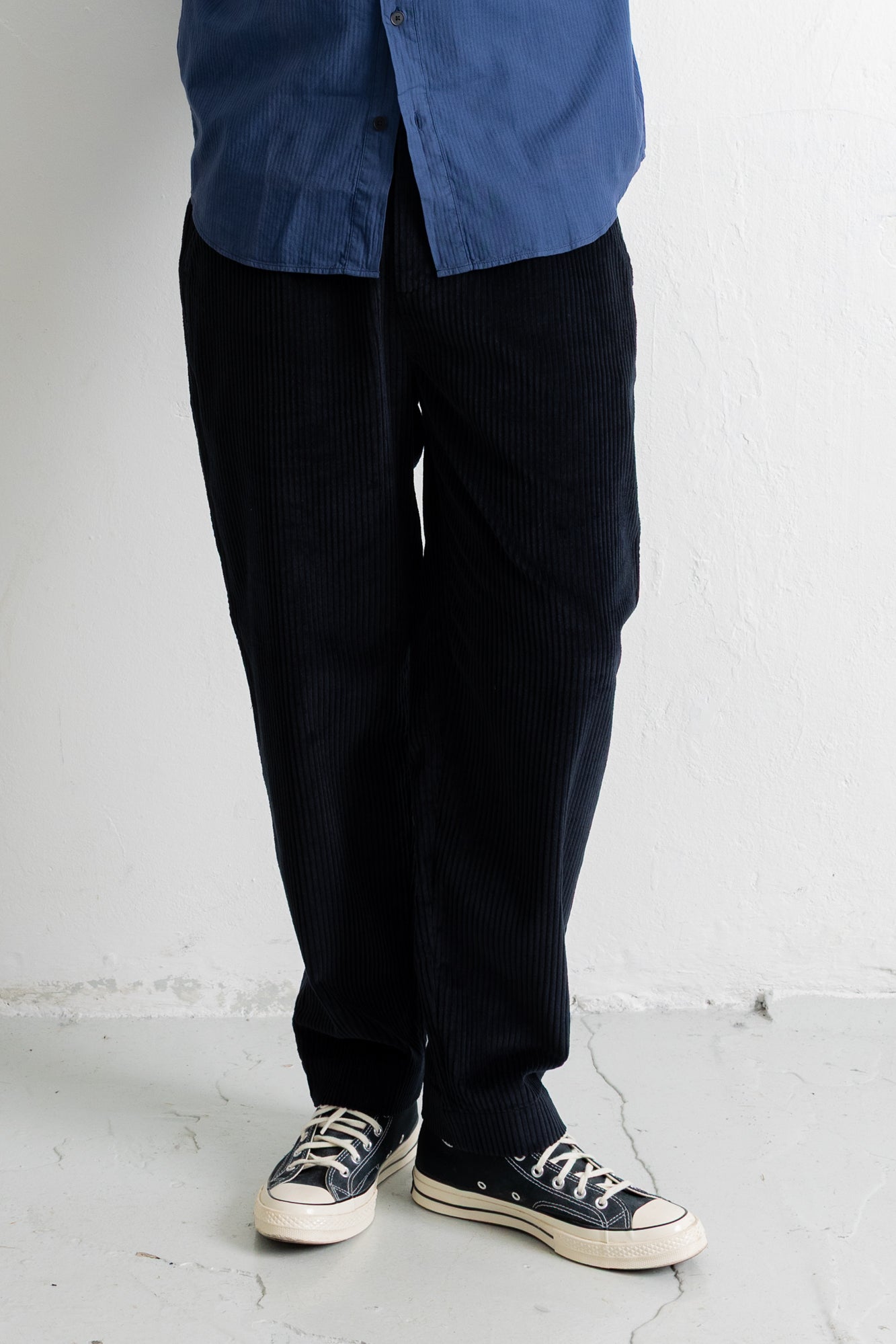 A person wearing dark navy corduroy pants, a blue button-up shirt, and black canvas sneakers, standing against a white wall.