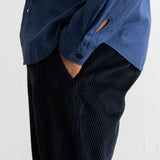 A close-up of a person wearing a blue button-up shirt and dark navy corduroy trousers, with one hand in a pocket, against a white textured wall.