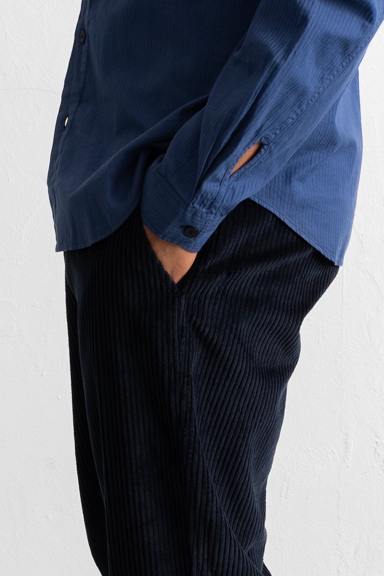 A close-up of a person wearing a blue button-up shirt and dark navy corduroy trousers, with one hand in a pocket, against a white textured wall.