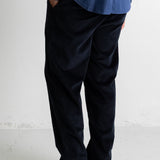 A person shown from the waist down wearing dark navy corduroy trousers, a blue shirt, and black canvas sneakers with white rubber soles, standing against a light gray wall.