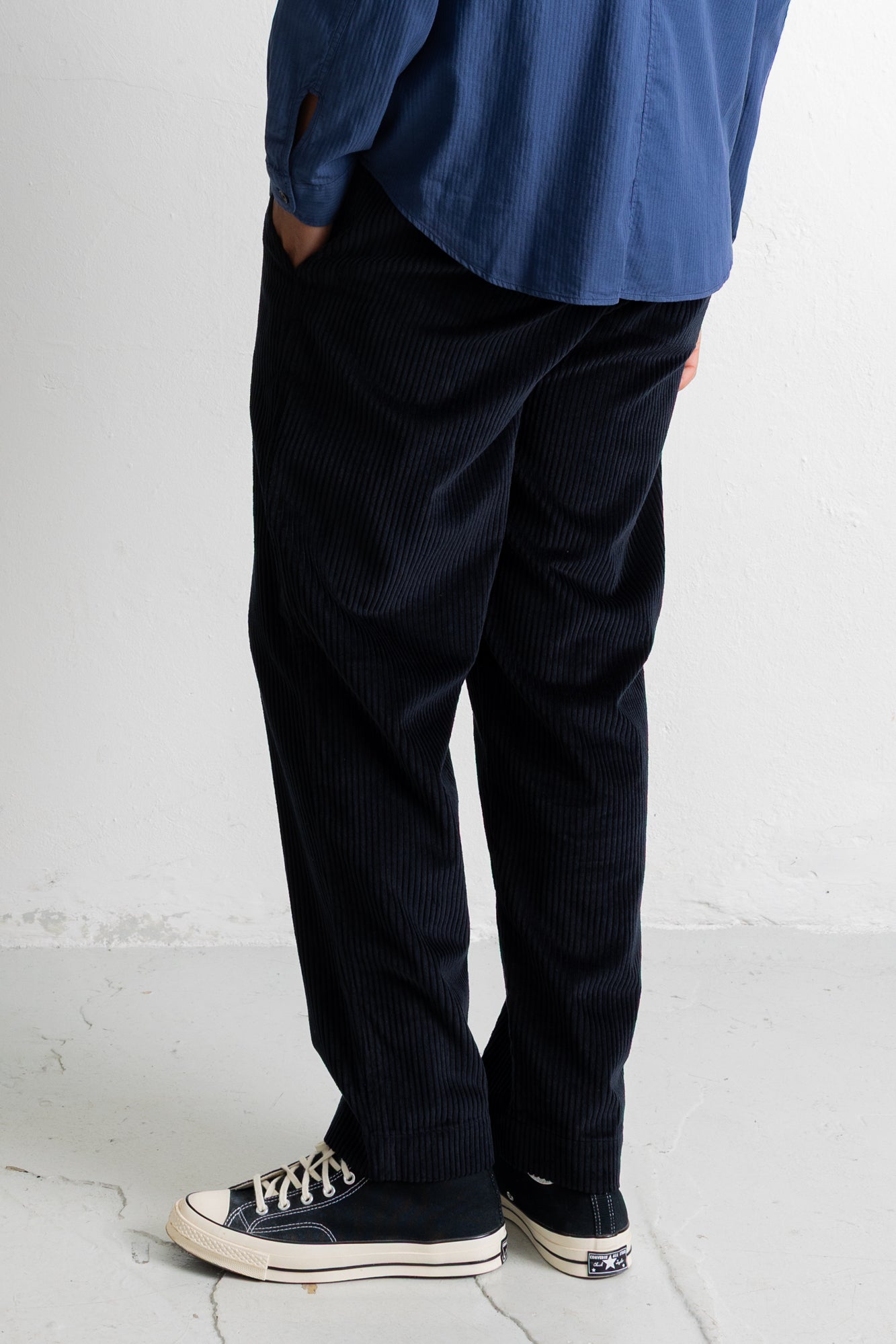 A person shown from the waist down wearing dark navy corduroy trousers, a blue shirt, and black canvas sneakers with white rubber soles, standing against a light gray wall.