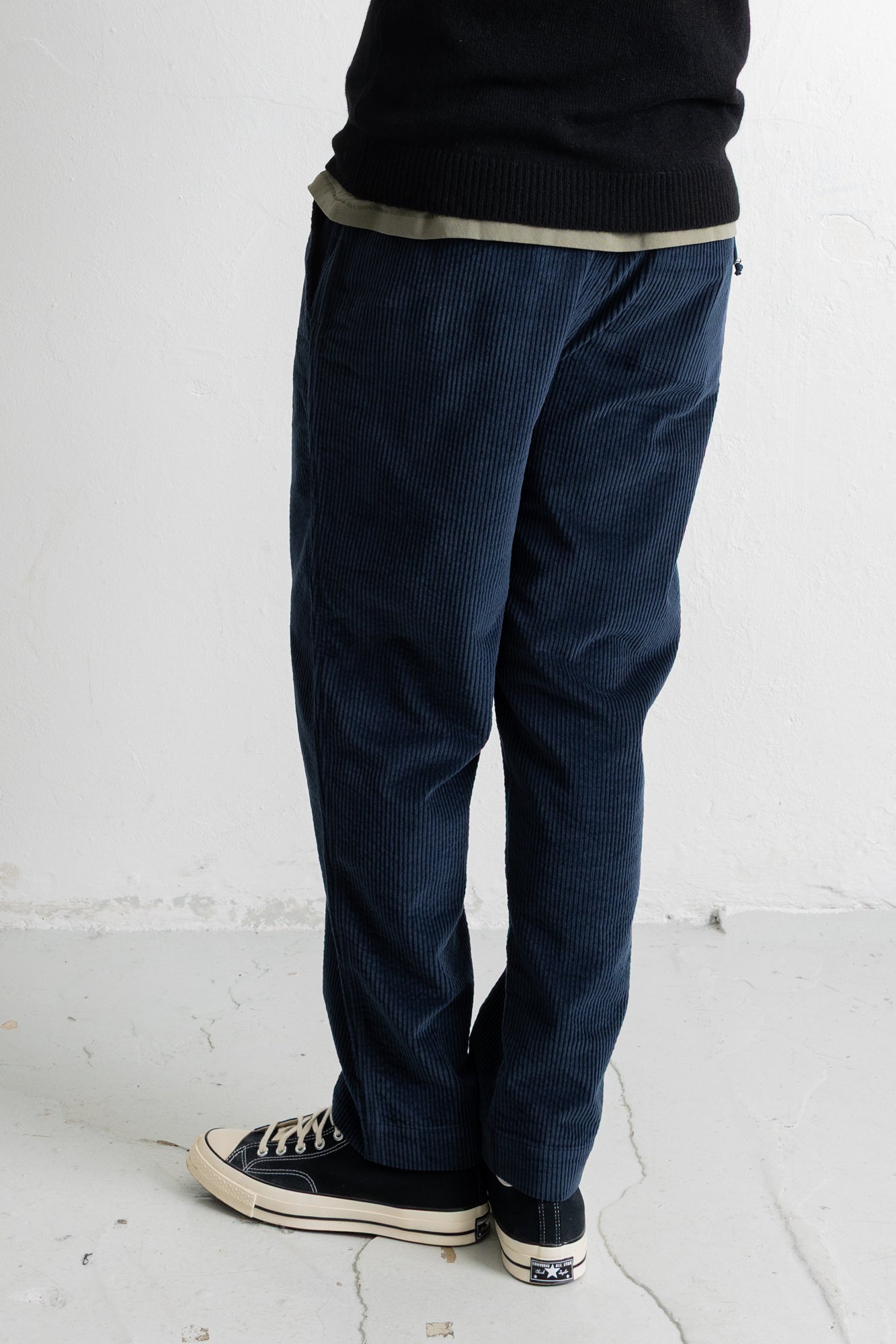 A person shown from the waist down wearing dark navy corduroy pants, a black knit top, and black canvas sneakers with white soles, standing against a white wall.