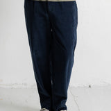 A person wearing dark navy corduroy trousers, a black sweater, and black canvas sneakers, standing against a white wall.