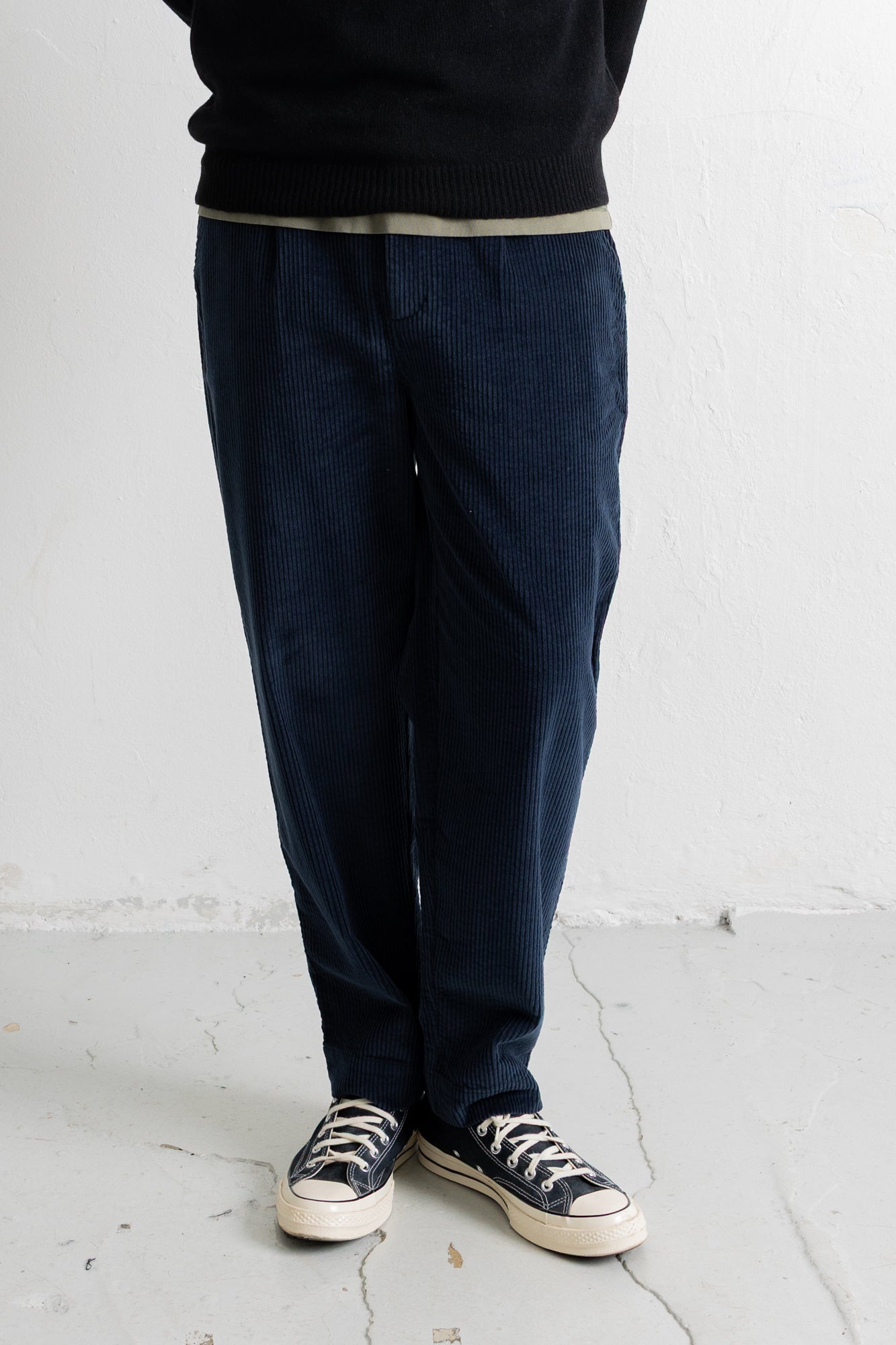 A person wearing dark navy corduroy trousers, a black sweater, and black canvas sneakers, standing against a white wall.