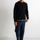 A man wearing a black crewneck sweatshirt, dark blue baggy pants, and black and white canvas sneakers, standing against a white studio wall.