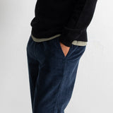A person wearing dark navy corduroy trousers and a black sweater, shown from the chest down against a plain light gray wall.
