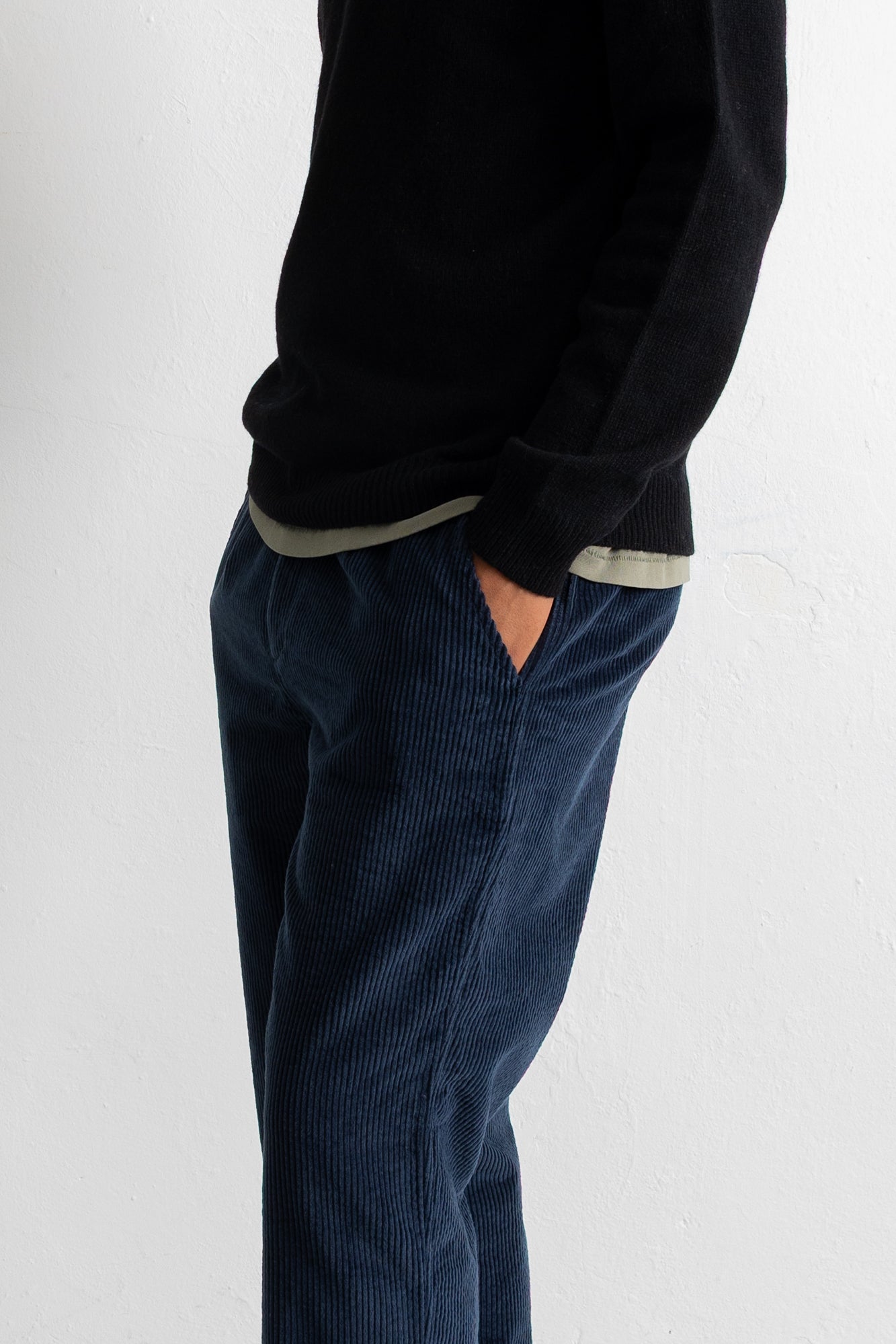 A person wearing dark navy corduroy trousers and a black sweater, shown from the chest down against a plain light gray wall.