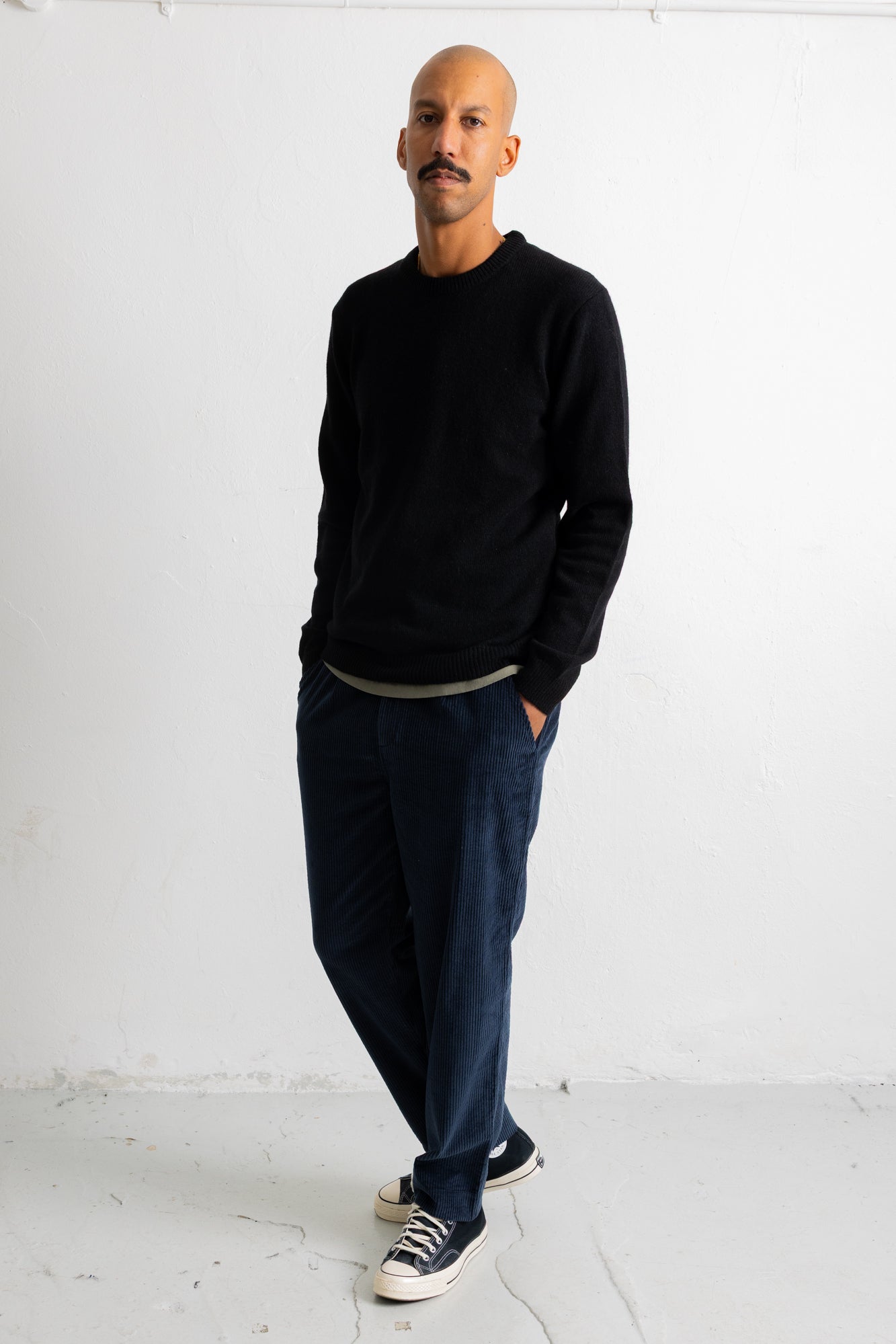 A man wearing a black crewneck sweater, dark blue jeans, and navy sneakers with white soles, standing against a white studio wall.