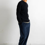 A man standing in profile wearing a black long-sleeve crewneck sweater, dark blue corduroy trousers, and black canvas sneakers against a plain white studio background.