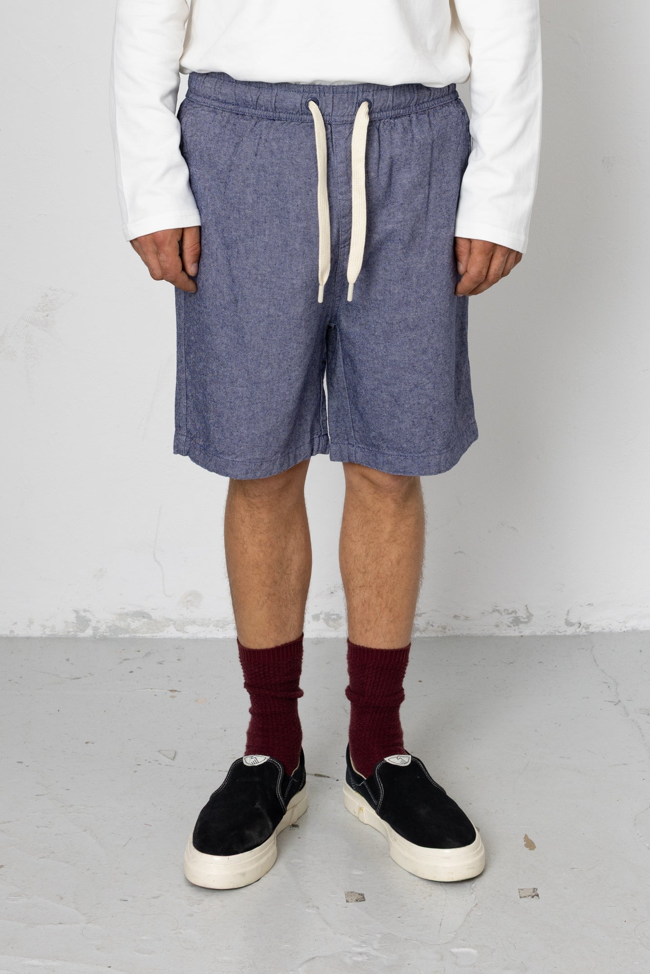 Person wearing blue shorts, white sweater, maroon socks, and black shoes on a plain background