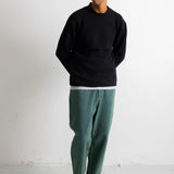 A man wearing a black crewneck sweatshirt, green relaxed-fit pants, a black baseball cap, and black canvas sneakers, standing against a white wall.