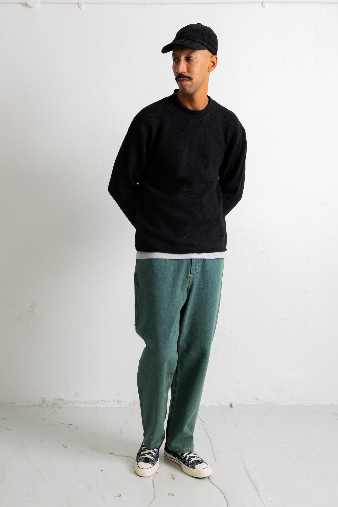 A man wearing a black crewneck sweatshirt, green relaxed-fit pants, a black baseball cap, and black canvas sneakers, standing against a white wall.