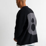 A man wearing a black knit sweater with a large gray circular graphic on the back, paired with a black baseball cap and teal jeans, shown from a side-back angle against a plain white wall.