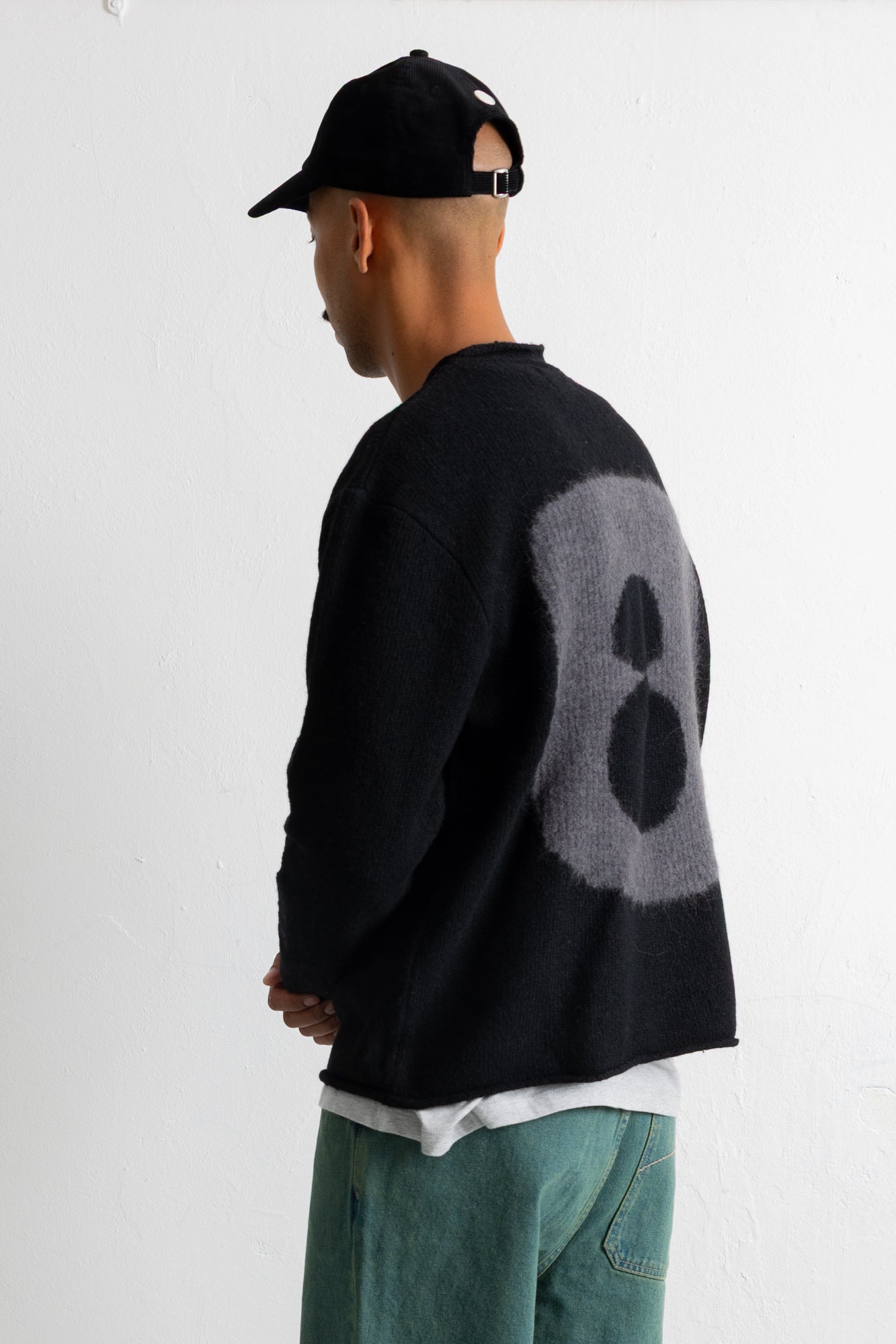 A man wearing a black knit sweater with a large gray circular graphic on the back, paired with a black baseball cap and teal jeans, shown from a side-back angle against a plain white wall.