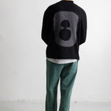 A man viewed from behind wearing a black knit sweater with a large gray number 8 on the back, green pants, and a black cap in a minimalist white studio setting.