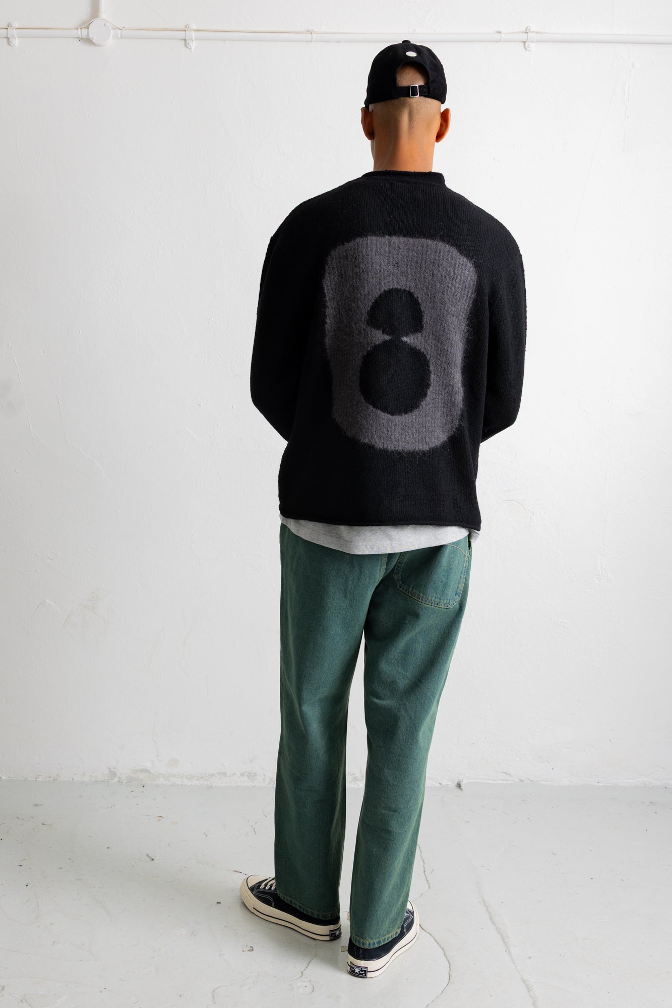 A man viewed from behind wearing a black knit sweater with a large gray number 8 on the back, green pants, and a black cap in a minimalist white studio setting.