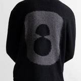 A person shown from the back wearing a black knit sweater with a large gray and black abstract circular graphic on the back, paired with teal pants and a light gray shirt underneath.