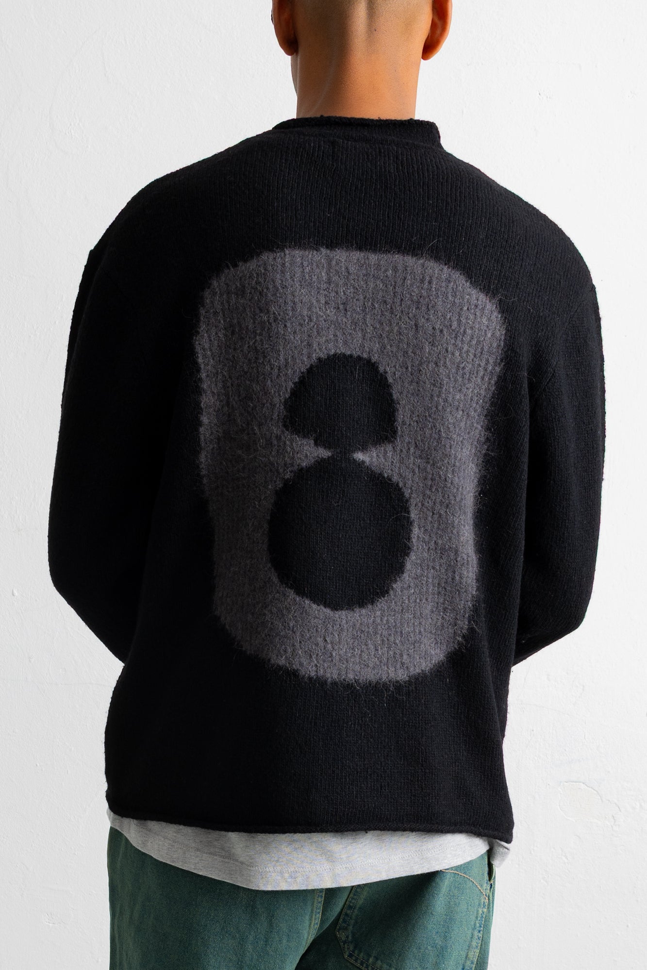 A person shown from the back wearing a black knit sweater with a large gray and black abstract circular graphic on the back, paired with teal pants and a light gray shirt underneath.