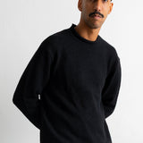 A man wearing a black crewneck sweater, a black baseball cap, and teal jeans, standing against a plain white wall.