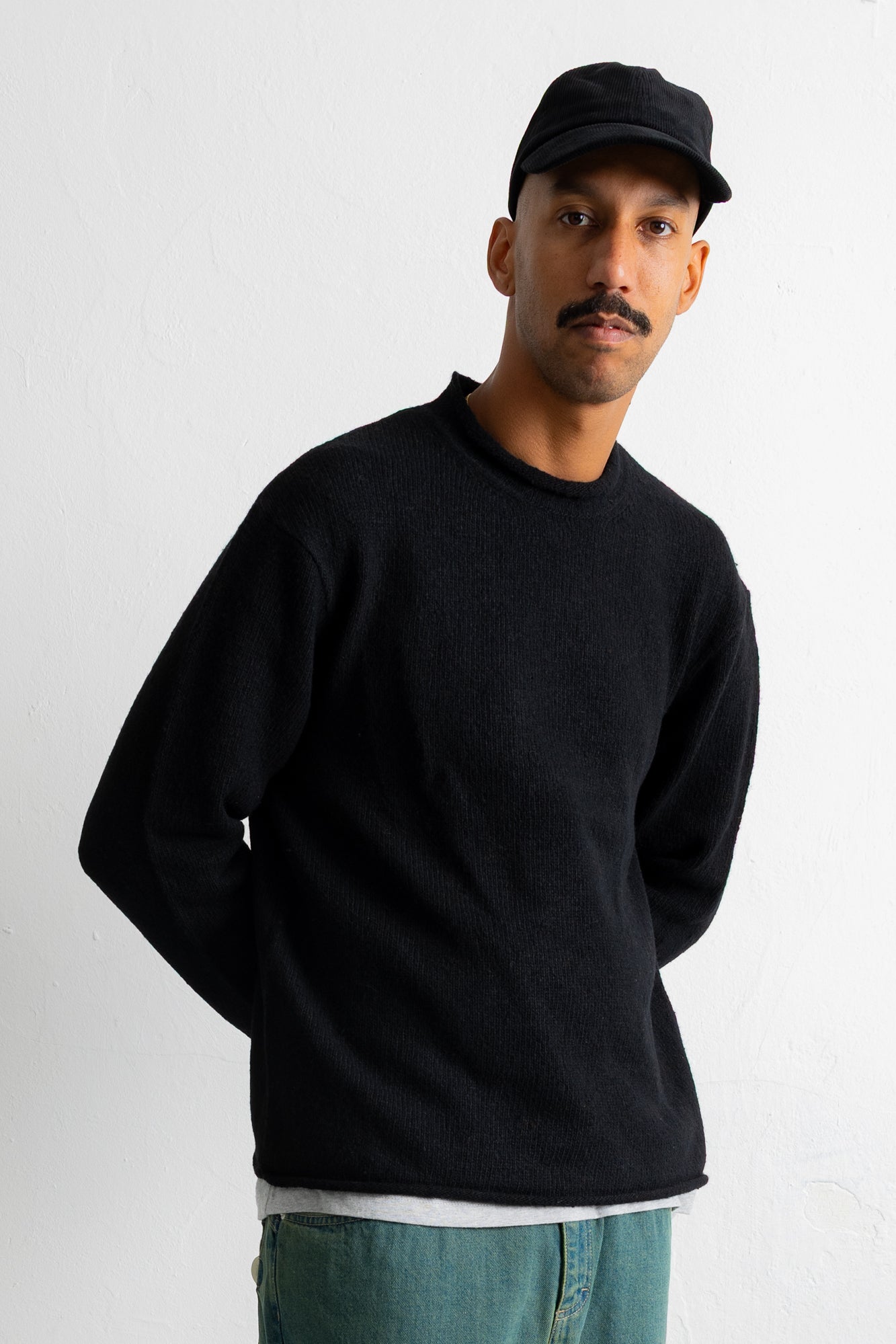 A man wearing a black crewneck sweater, a black baseball cap, and teal jeans, standing against a plain white wall.