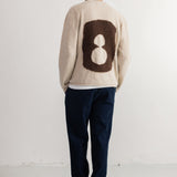 A man viewed from behind wearing a cream-colored knit sweater with a large brown and cream number 8 motif on the back, paired with dark navy pants and black shoes, standing against a white studio wall.