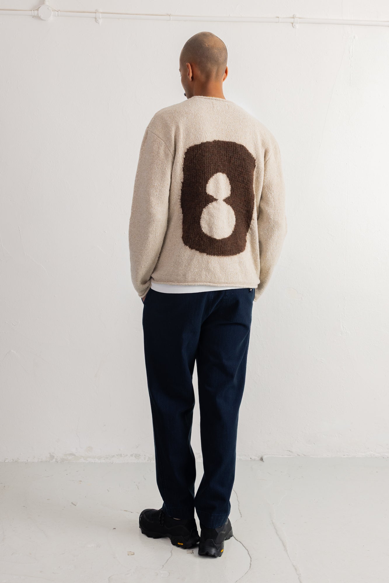 A man viewed from behind wearing a cream-colored knit sweater with a large brown and cream number 8 motif on the back, paired with dark navy pants and black shoes, standing against a white studio wall.